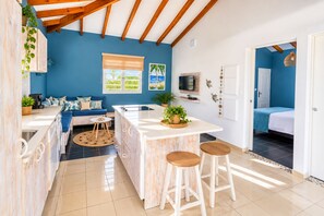 Een compacte keuken met een centraal eiland, een eethoek met krukken, een bank en een aan de muur bevestigde televisie.