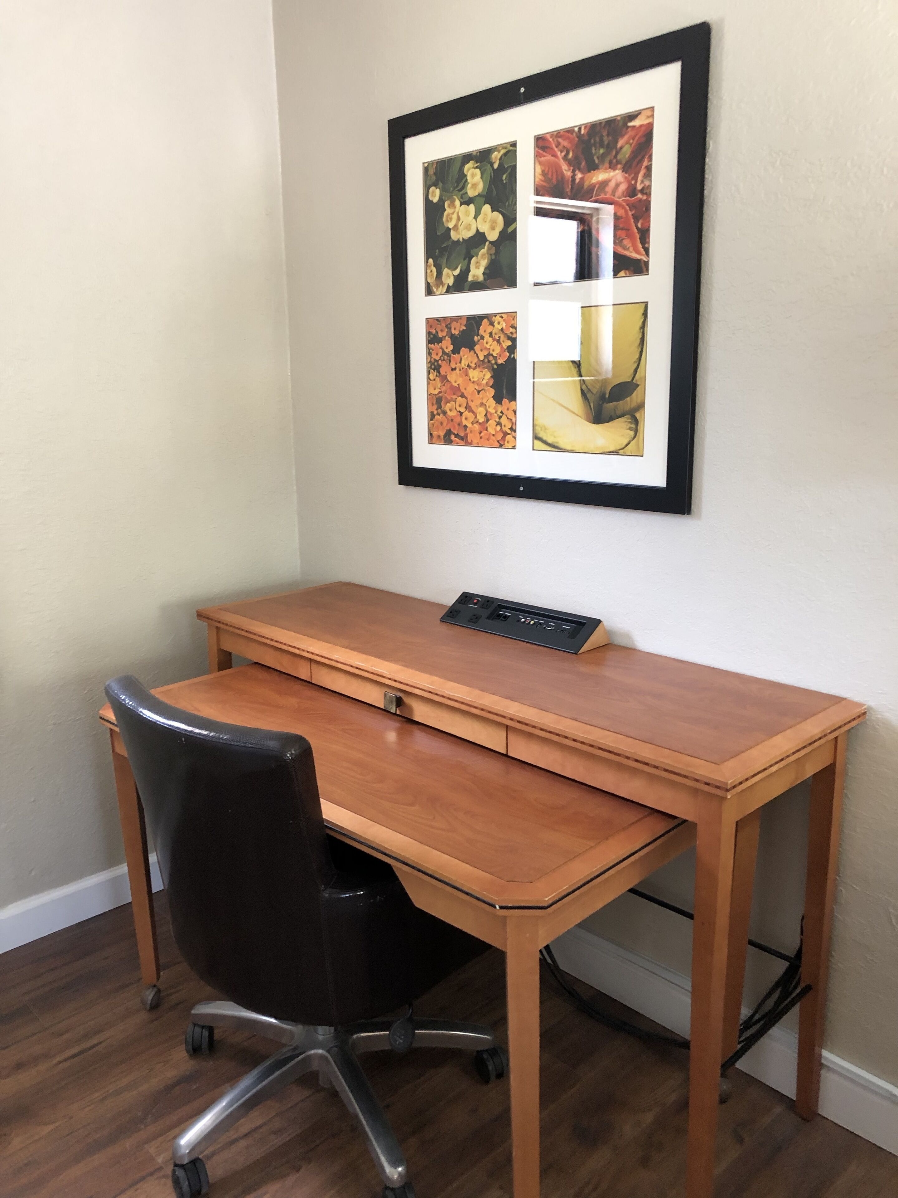 A wooden desk with a black office chair, a framed picture on the wall, and a power strip on the desk.