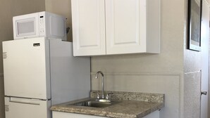 A compact kitchen with white cabinetry, a built-in microwave, a stainless steel sink, and a trash can. - Ocala Cove Motel (Ocala)