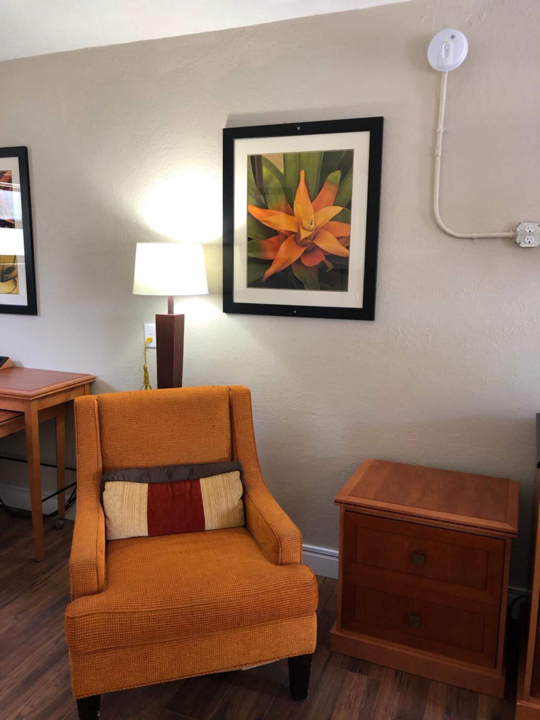 Ein Zimmer mit einem einzelnen orangefarbenen Sessel, einem hölzernen Nachttisch, einem gerahmten Bild eines Blütenzweigs an der Wand und einer Lampe auf einem Beistelltisch.