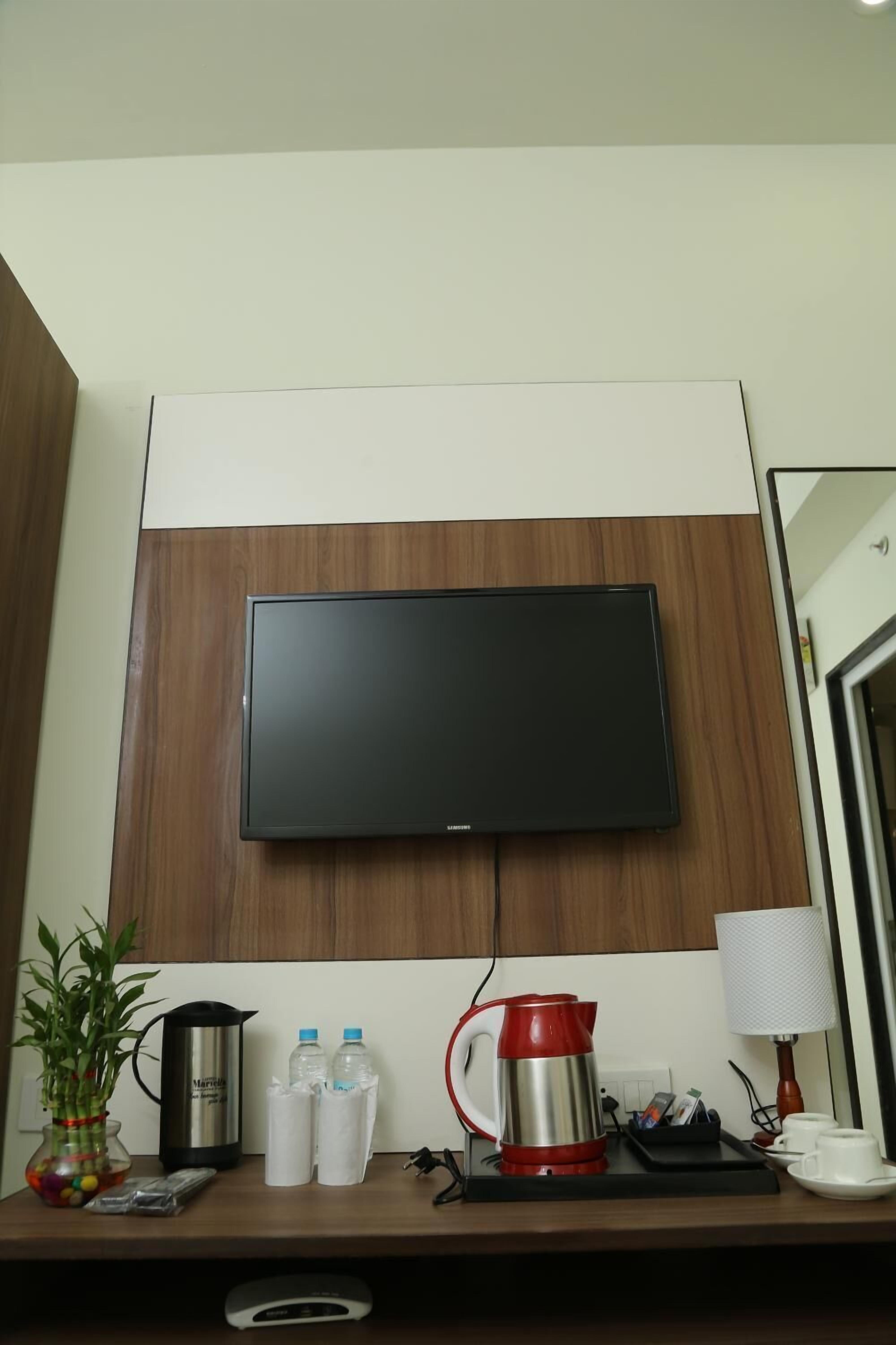 Habitación de hotel con televisor de pantalla plana montado en una pared de madera, cafetera, dos botellas de agua, hervidor rojo, lámpara y una mesita con un jarrón de flores.