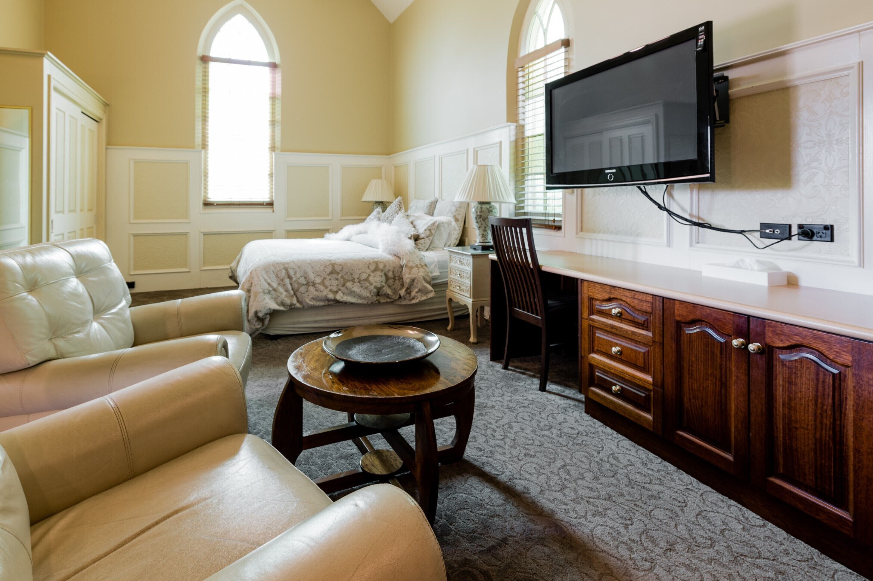 A hotel room with a bed, a sofa, a dining table, and a TV mounted on the wall.