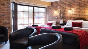 A hotel room with a brick wall, a large window, two beds with red and white bedding, black leather chairs, and a hanging light fixture.