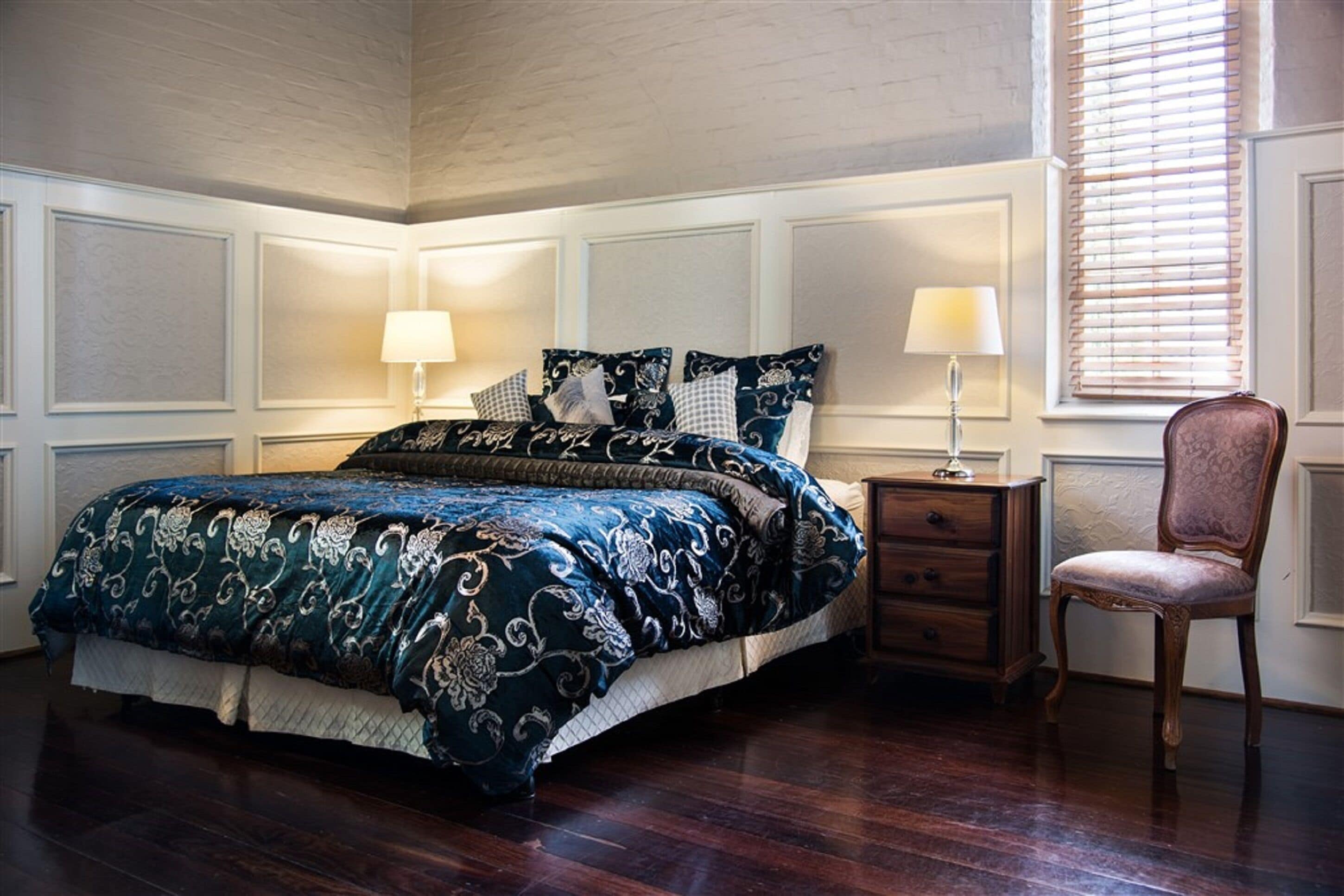 A bedroom with a large bed, two bedside tables, a chair, and a window with blinds.