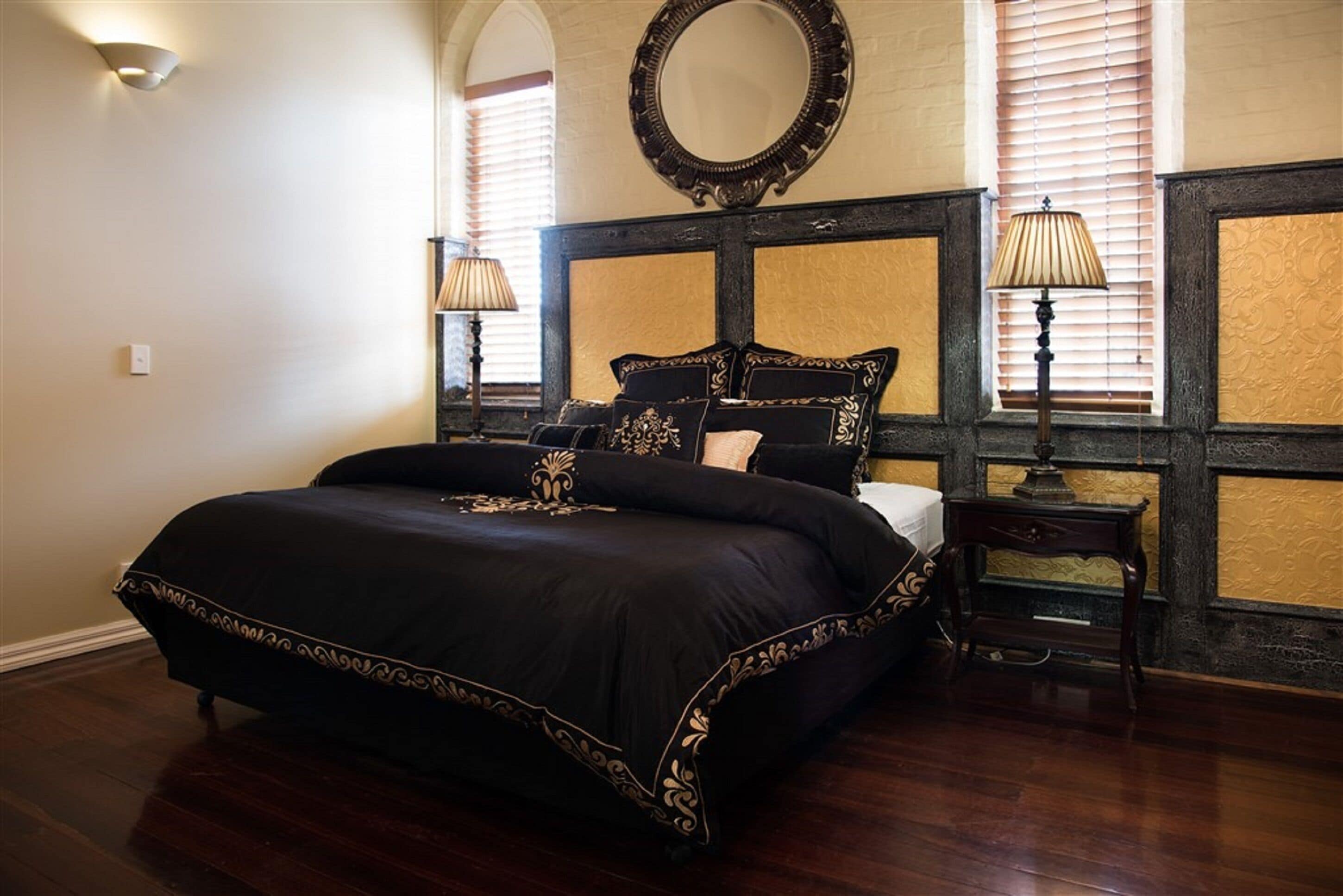 A bedroom with a large bed, two bedside tables, a round mirror above the bed, and a window with blinds.