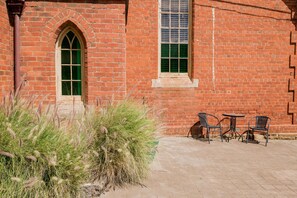 A brick building with a large arched window and a smaller window with shutters. There are outdoor tables and chairs.