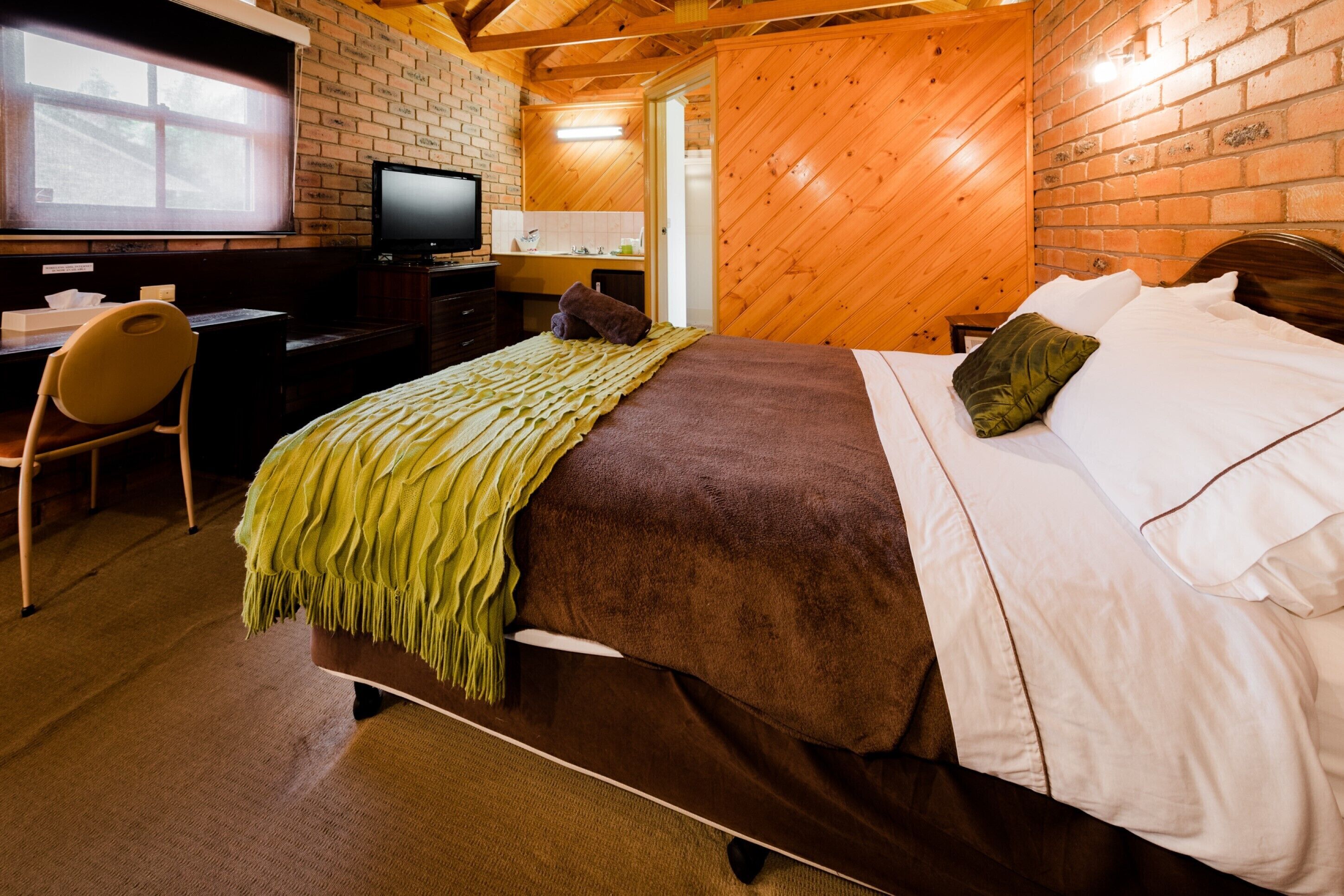 A bedroom with a bed, a desk, a chair, a television, and a brick wall.