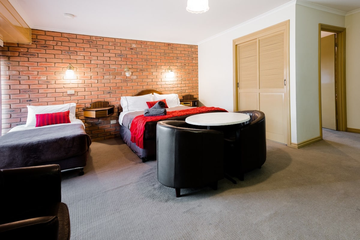 A hotel room with a brick wall, a bed with red bedding, a small table, two chairs, and a wardrobe.