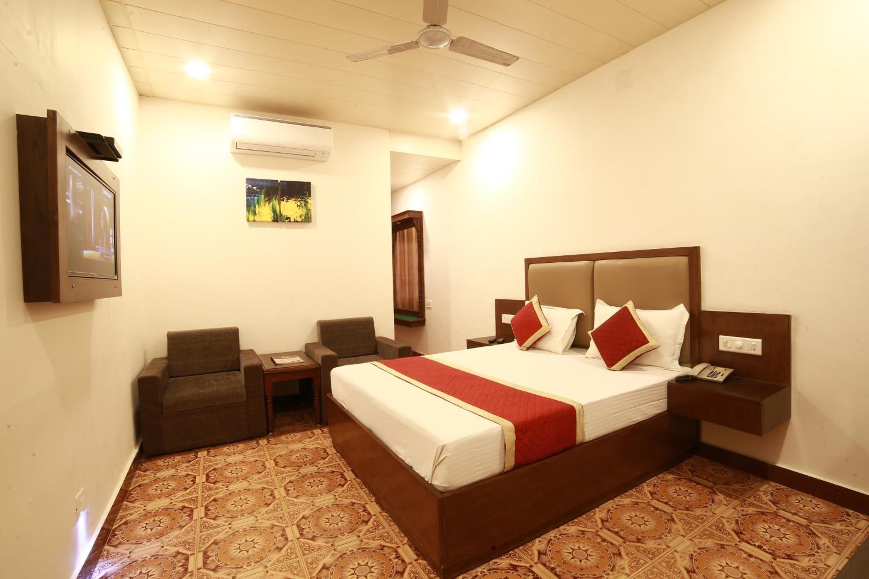 A hotel room with a large bed, two bedside tables, a chair, an air conditioning unit, a ceiling fan, and a wall-mounted TV.