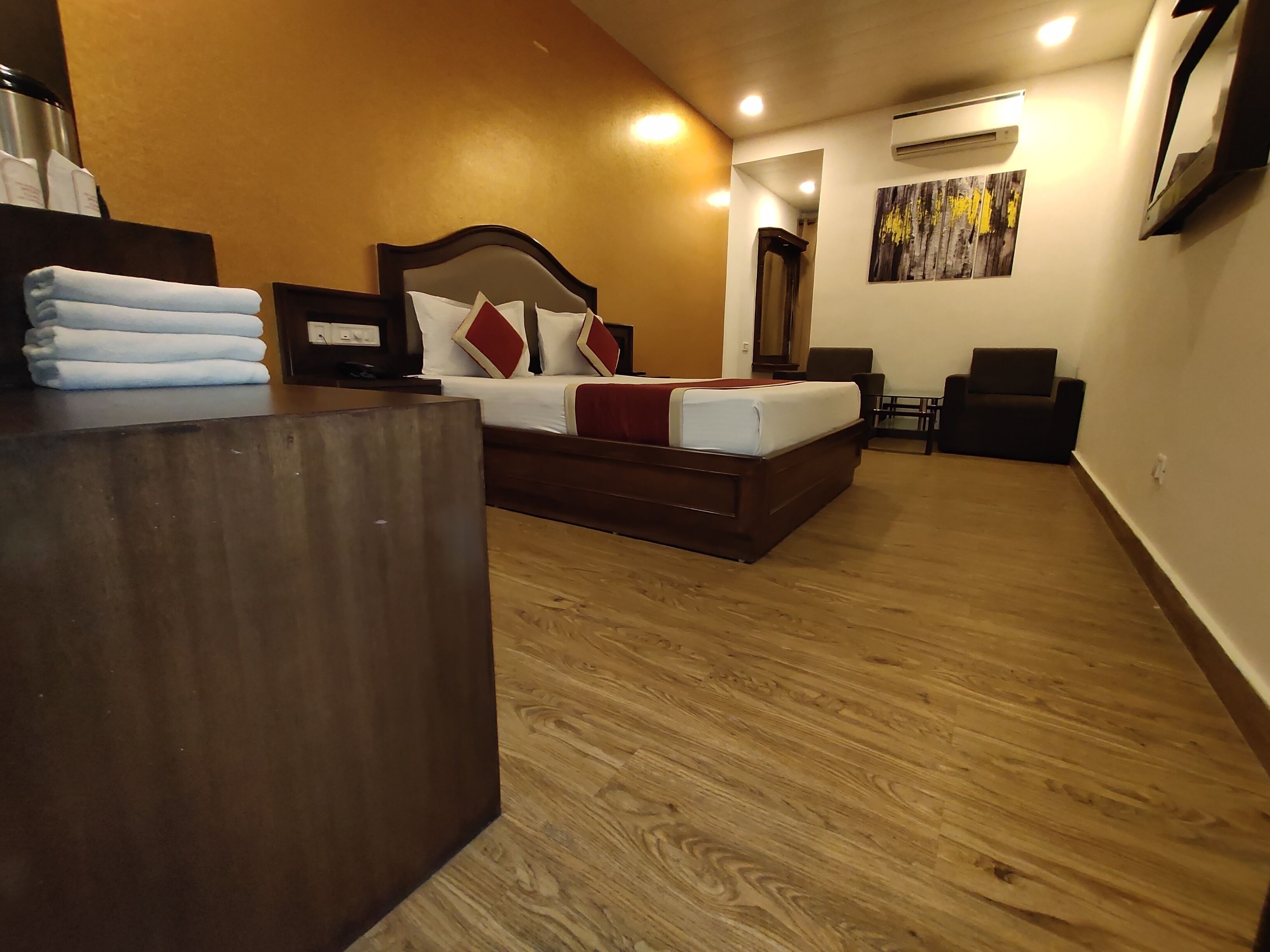 Quarto de hotel com piso de madeira, cama com roupa de cama branca e almofadas vermelhas, escrivaninha com uma pilha de toalhas e unidade de ar condicionado na parede.