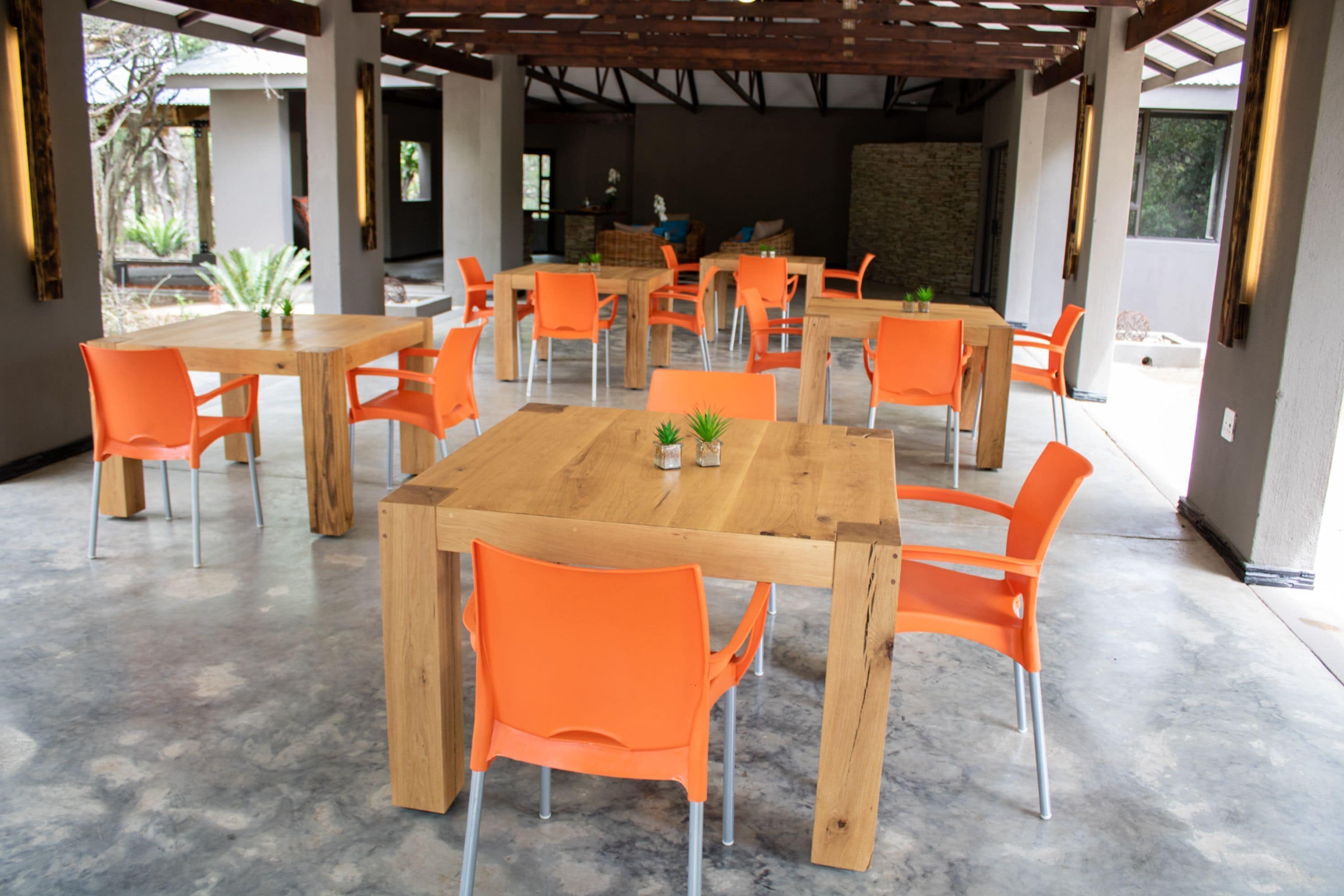 A spacious outdoor dining area with wooden tables and orange chairs, featuring a stone wall and large windows.