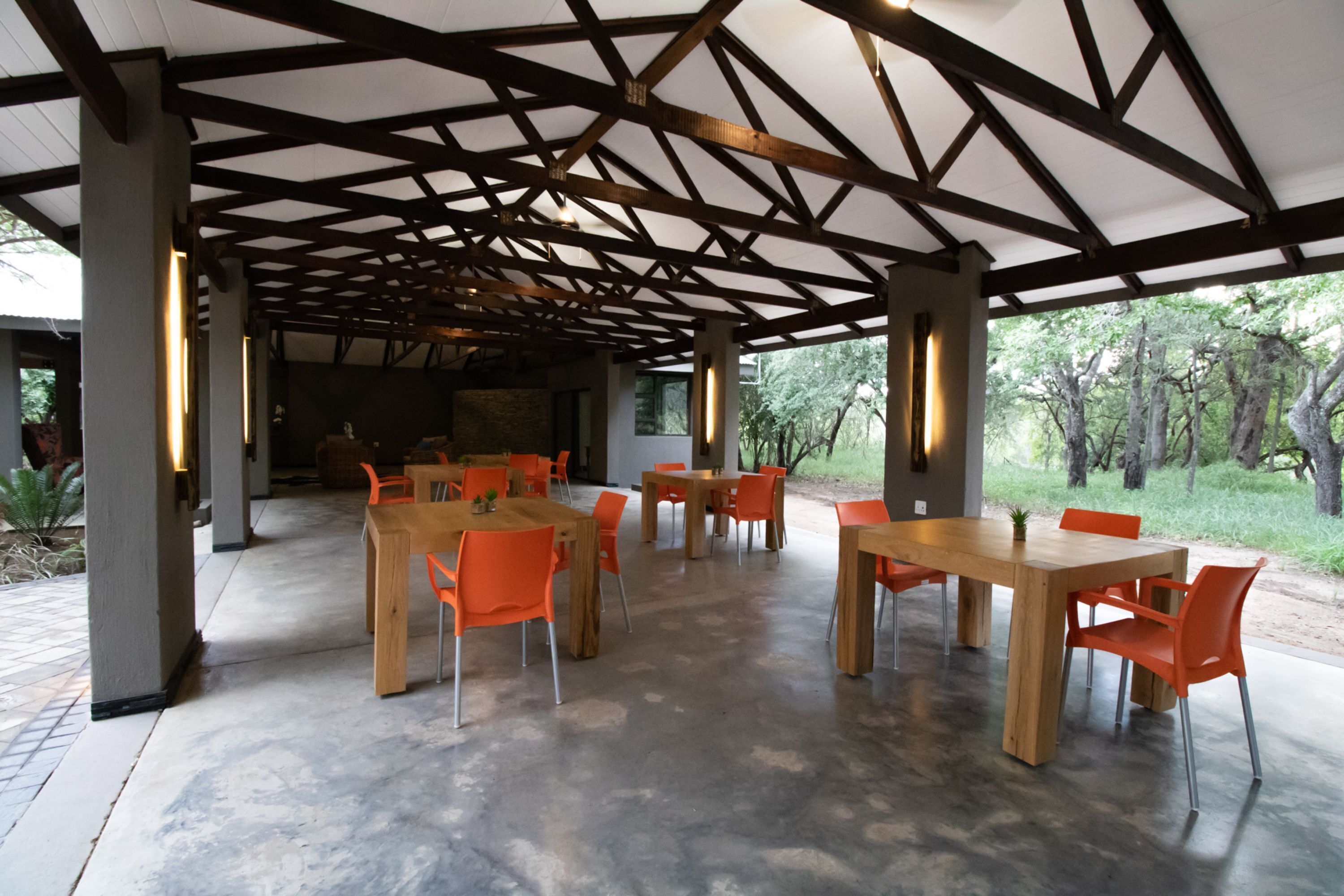 An open-plan dining area with wooden tables and orange chairs, under a wooden beam roof structure.