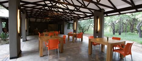 An open-plan dining area with wooden tables and orange chairs, under a wooden beam roof structure.