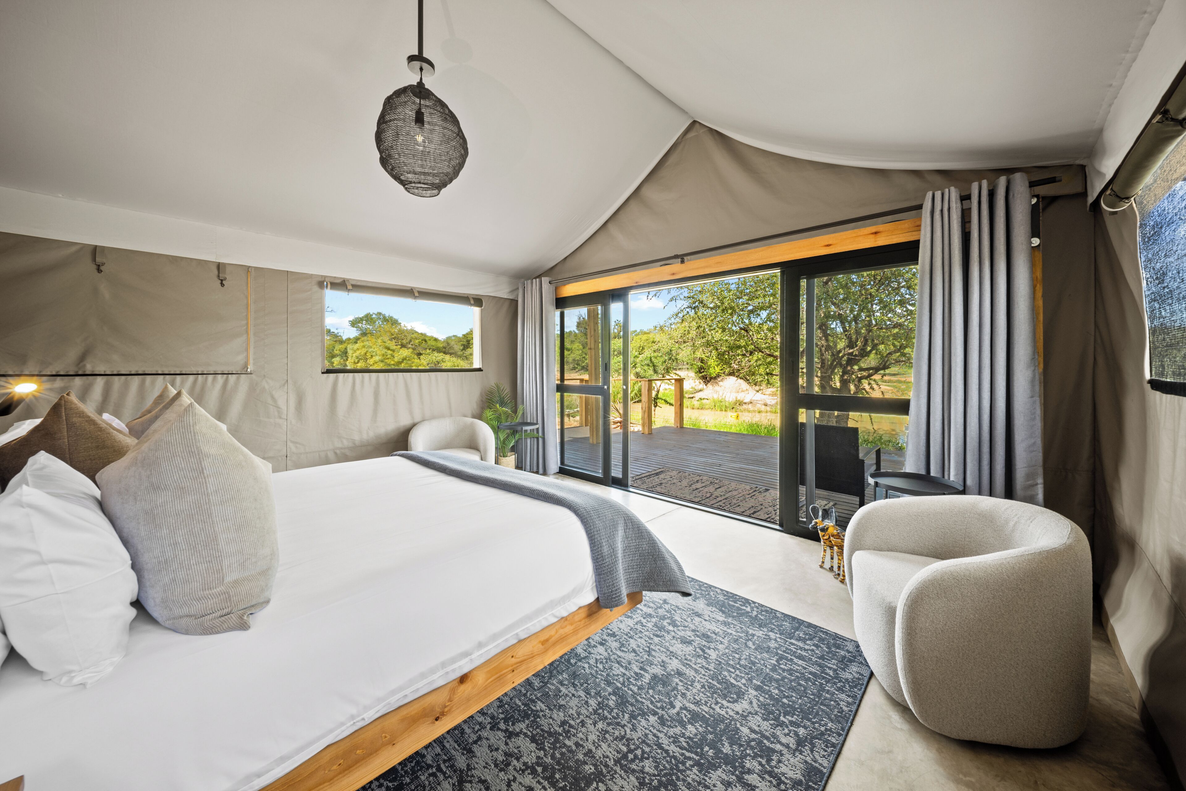 A bedroom with a large bed, a chair, a hanging light fixture, and a view through a glass door.
