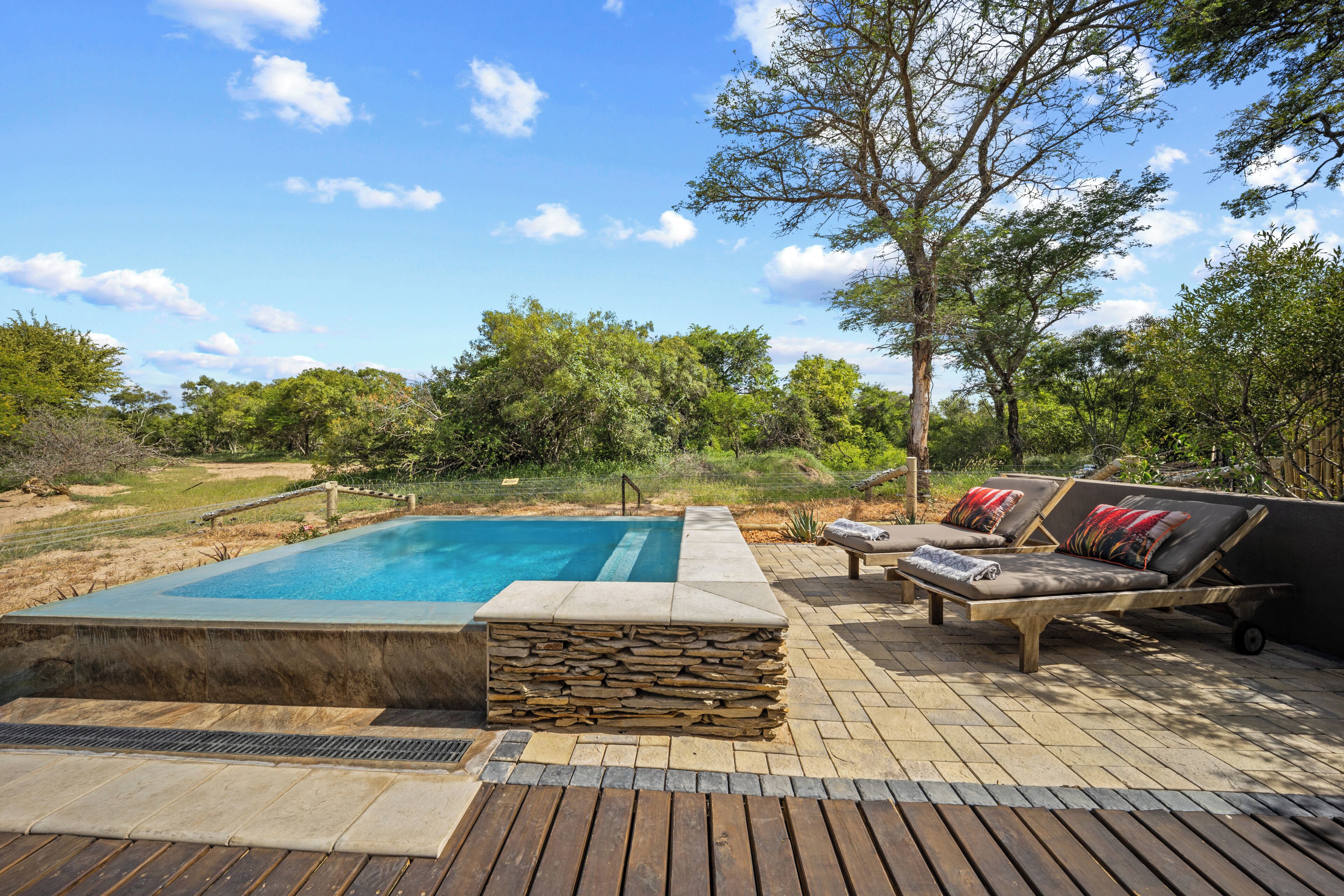 Une piscine entourée de pierres, avec une terrasse en bois et des chaises longues dotées de coussins.