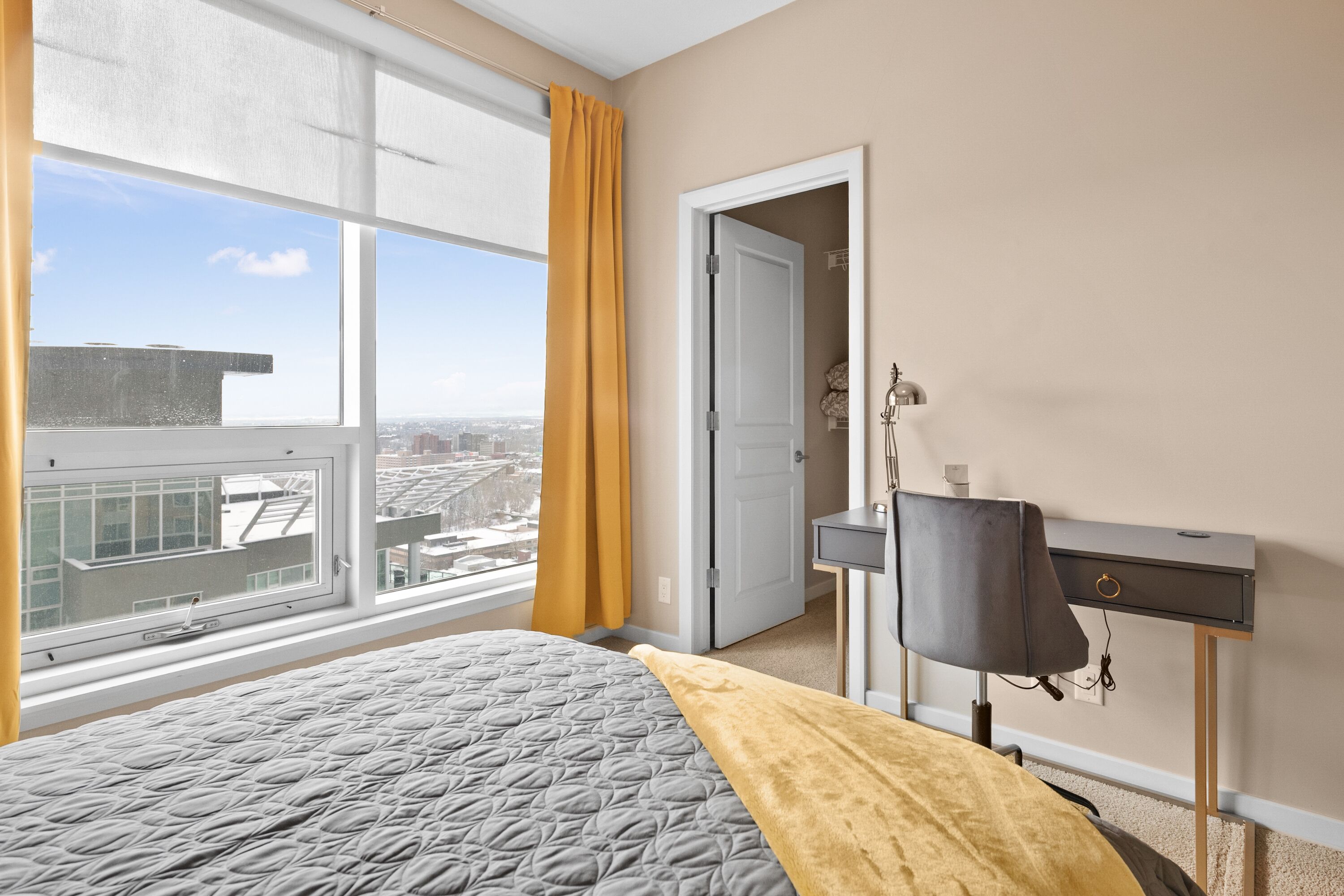 A hotel room with a large window, a bed with a patterned bedspread, a desk with a lamp, and a view of the cityscape.
