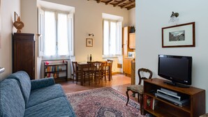Een woonkamer met een bank, een televisie, een boekenkast, een eethoek met stoelen en een tafel, en een keuken op de achtergrond.