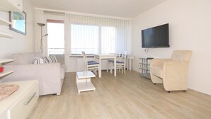 Ein modernes, gut beleuchtetes Wohnzimmer mit einer Couch, einem Esstisch, Stühlen und einem an der Wand befestigten Fernseher.