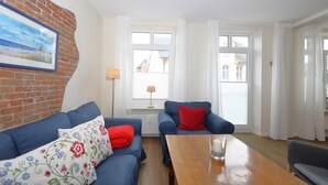 A living room with a blue sofa, a wooden coffee table, and a red cushion. There is a painting on the wall and a window with curtains. - Möwennest, Germany (Sylt)