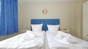 A double bed with white linens and two pillows, a blue headboard, a bedside table with a lamp, and a blue curtain. - Möwennest, Germany (Sylt)