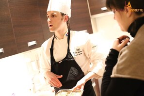 Um cozinheiro de uniforme branco e avental preto, em uma cozinha.