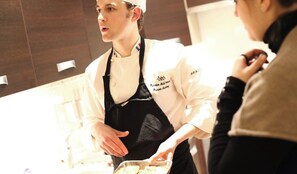 Een kok in een wit uniform met een zwarte schort, staand in een keuken.