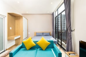 A modern hotel room with a large bed, a sofa with yellow pillows, a wooden panel wall, and a view of the city through large windows.
