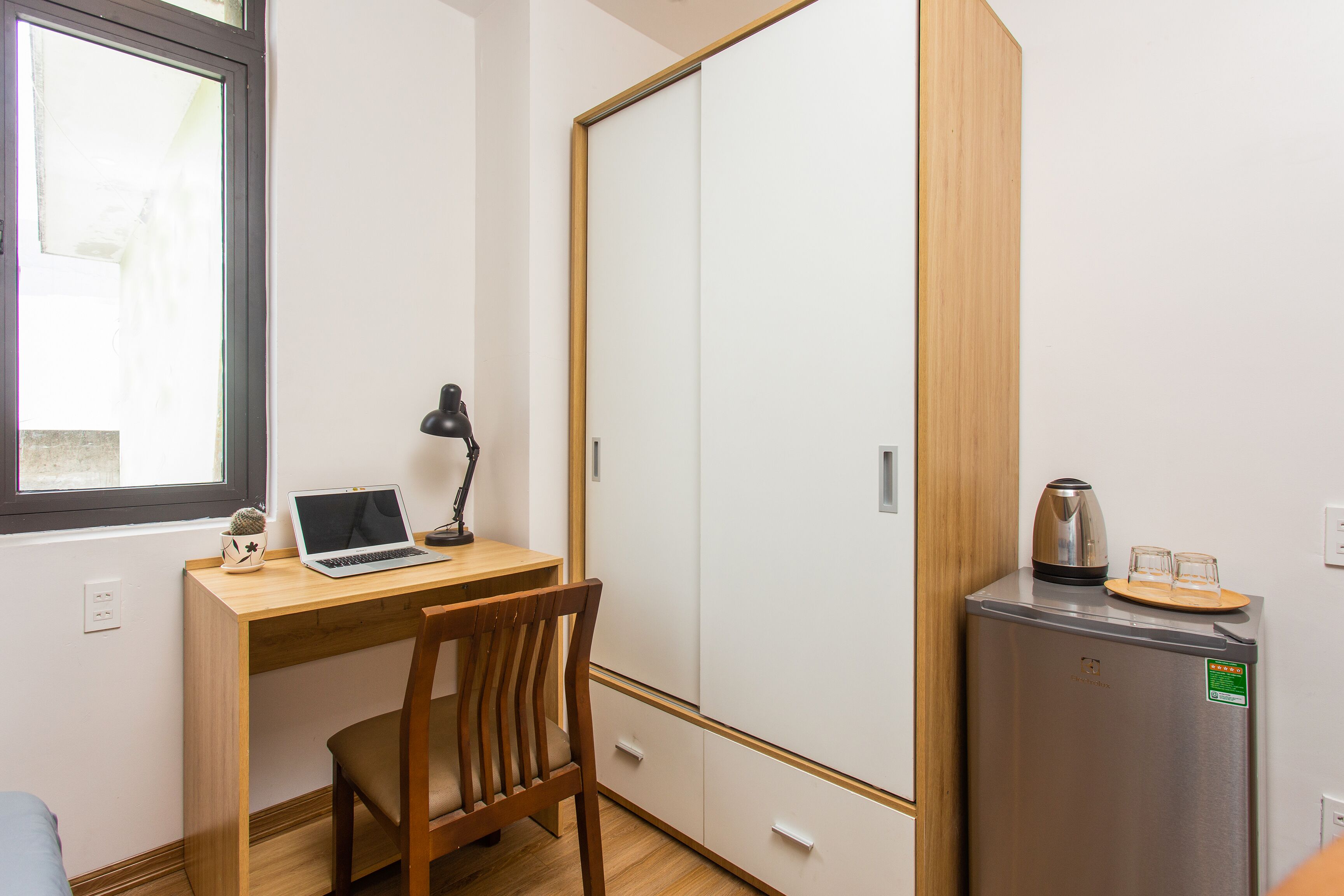 Una habitación compacta con escritorio de madera, una computadora portátil, una silla, un refrigerador pequeño y un armario con puerta corrediza.