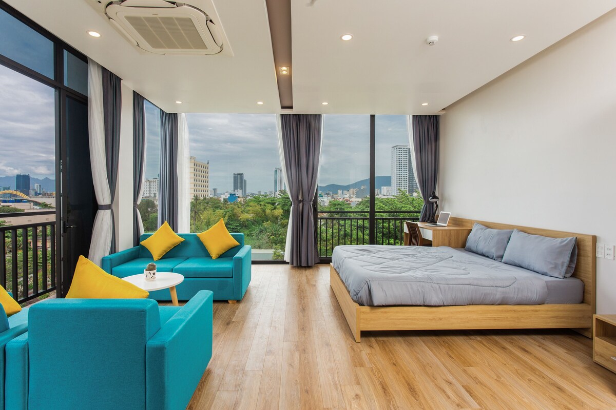 A modern hotel room with a large bed, a sofa, and a small table. There are large windows offering a city view.
