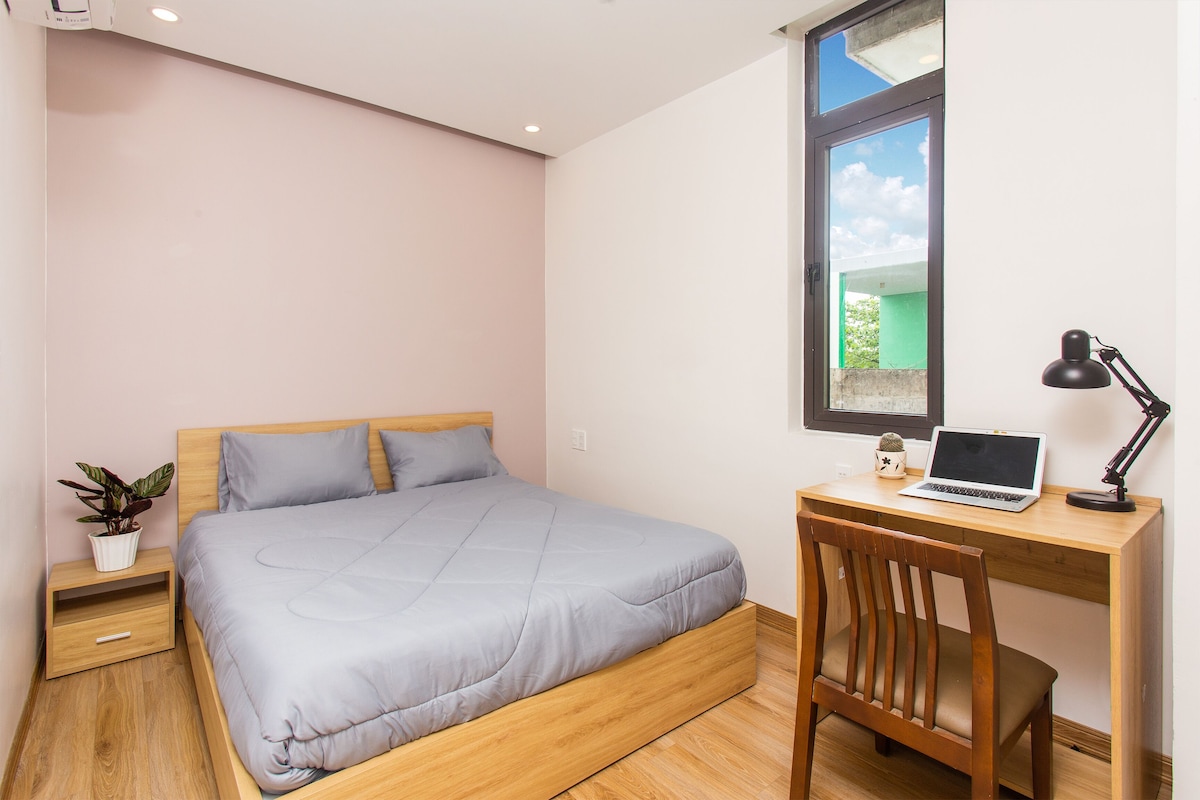 A bedroom with a bed, a desk, a laptop, a lamp, and a potted plant.