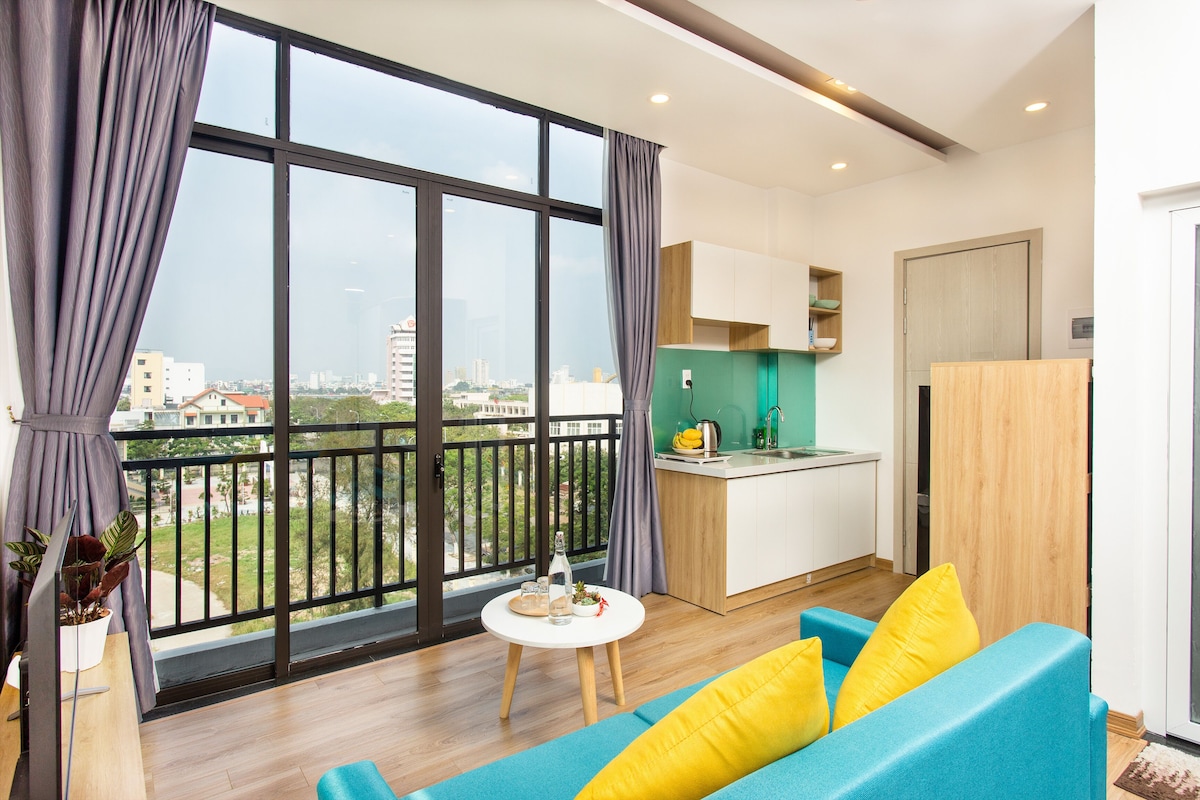 A modern apartment with a balcony, a kitchen, and a living area with a blue sofa and yellow cushions.