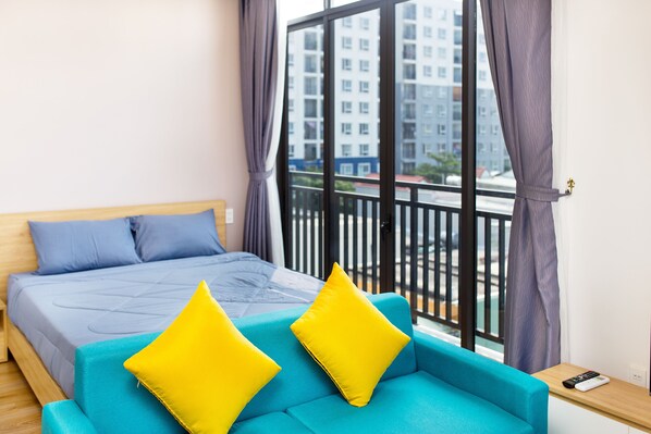 A bedroom with a bed, a blue sofa with yellow cushions, and a view of a building through a window with curtains.