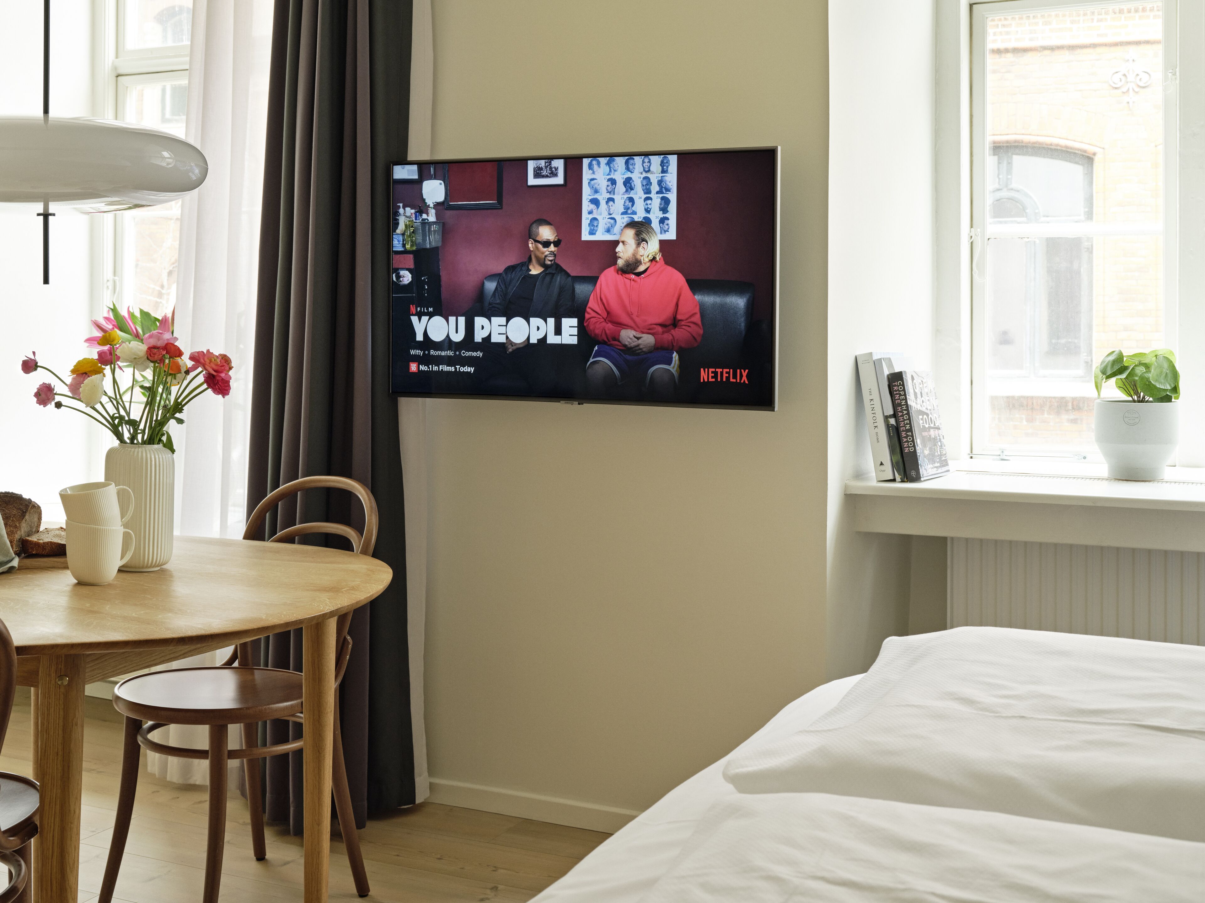 A hotel room with a bed, a TV mounted on the wall displaying a Netflix show, a dining table with chairs, a vase with flowers, and a window with a view of the outside.