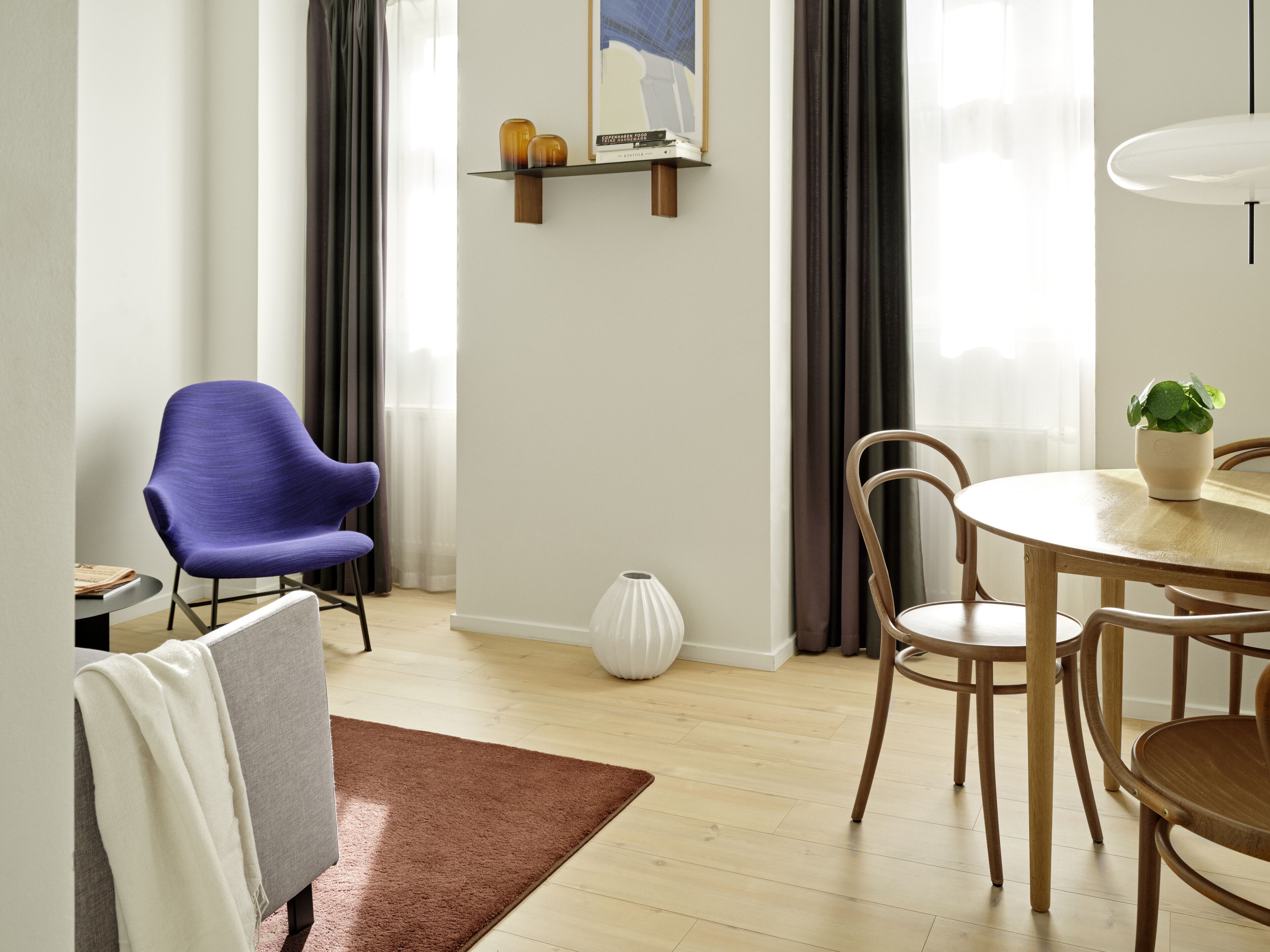 A modern living room with a grey sofa, a blue armchair, a round wooden dining table with chairs, a white vase, and a small shelf with books and decorative items.