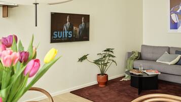 Una sala de estar moderna con un sofá gris, una mesa de centro, una planta en maceta y un televisor de pared que muestra 'SUITS'.