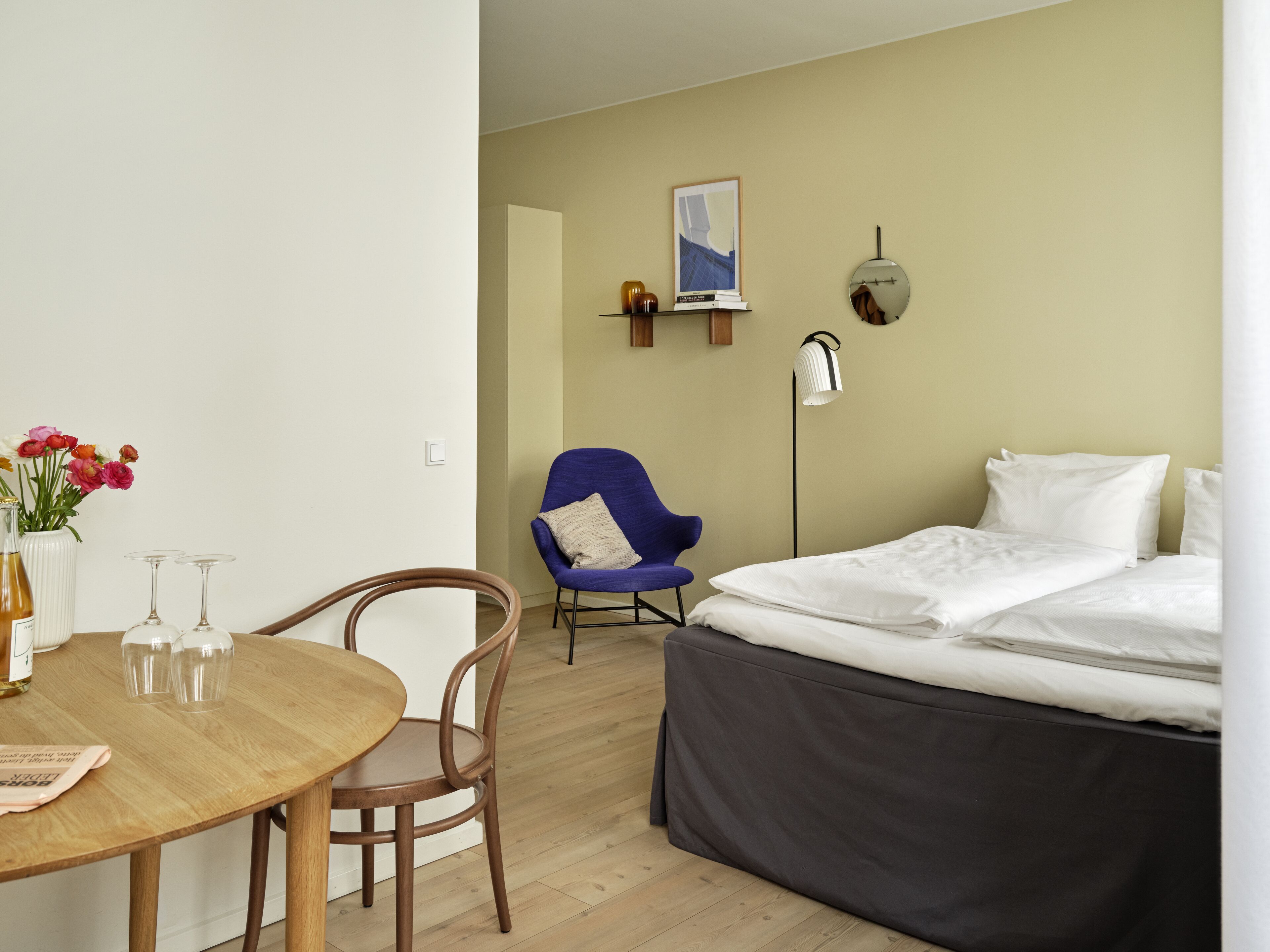 A hotel room with a bed, a chair, a table with a vase of flowers, and a wall-mounted shelf with books and a picture.