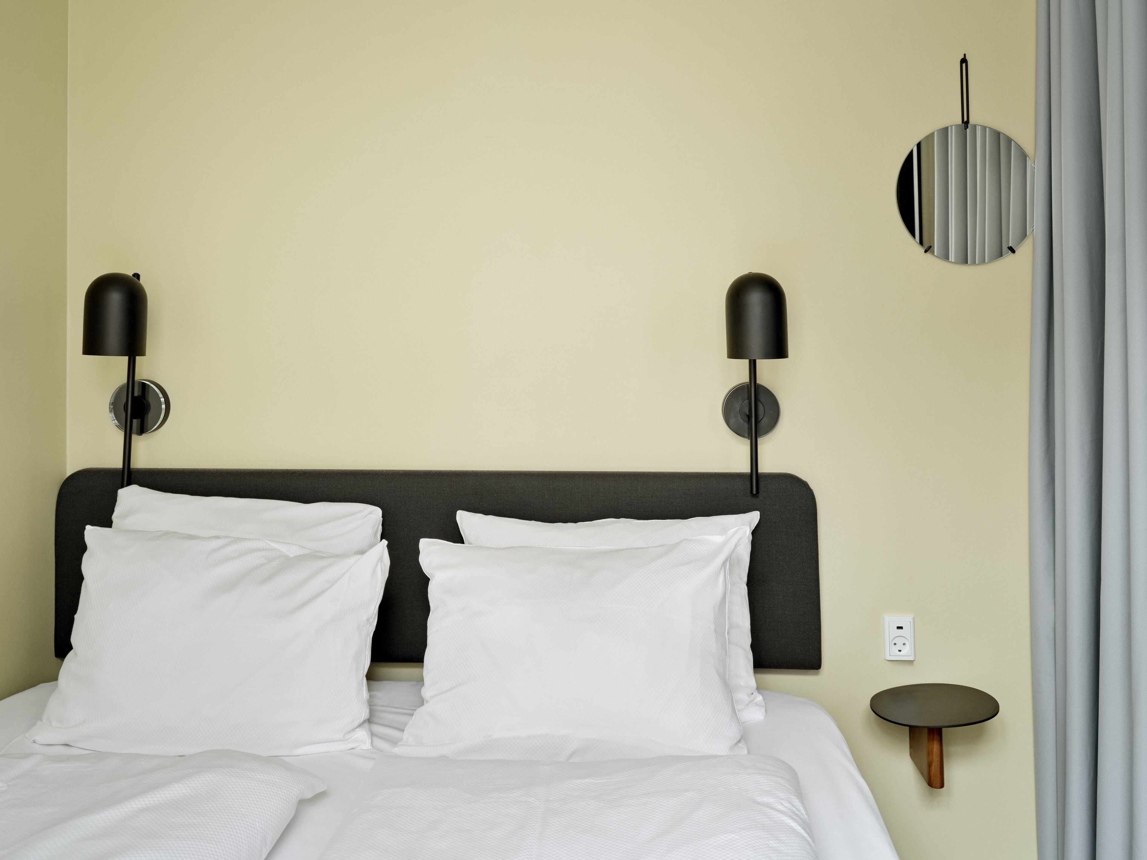 A neatly made bed with white linens, two wall-mounted black lamps, a round mirror, and a small round table with a wooden base.