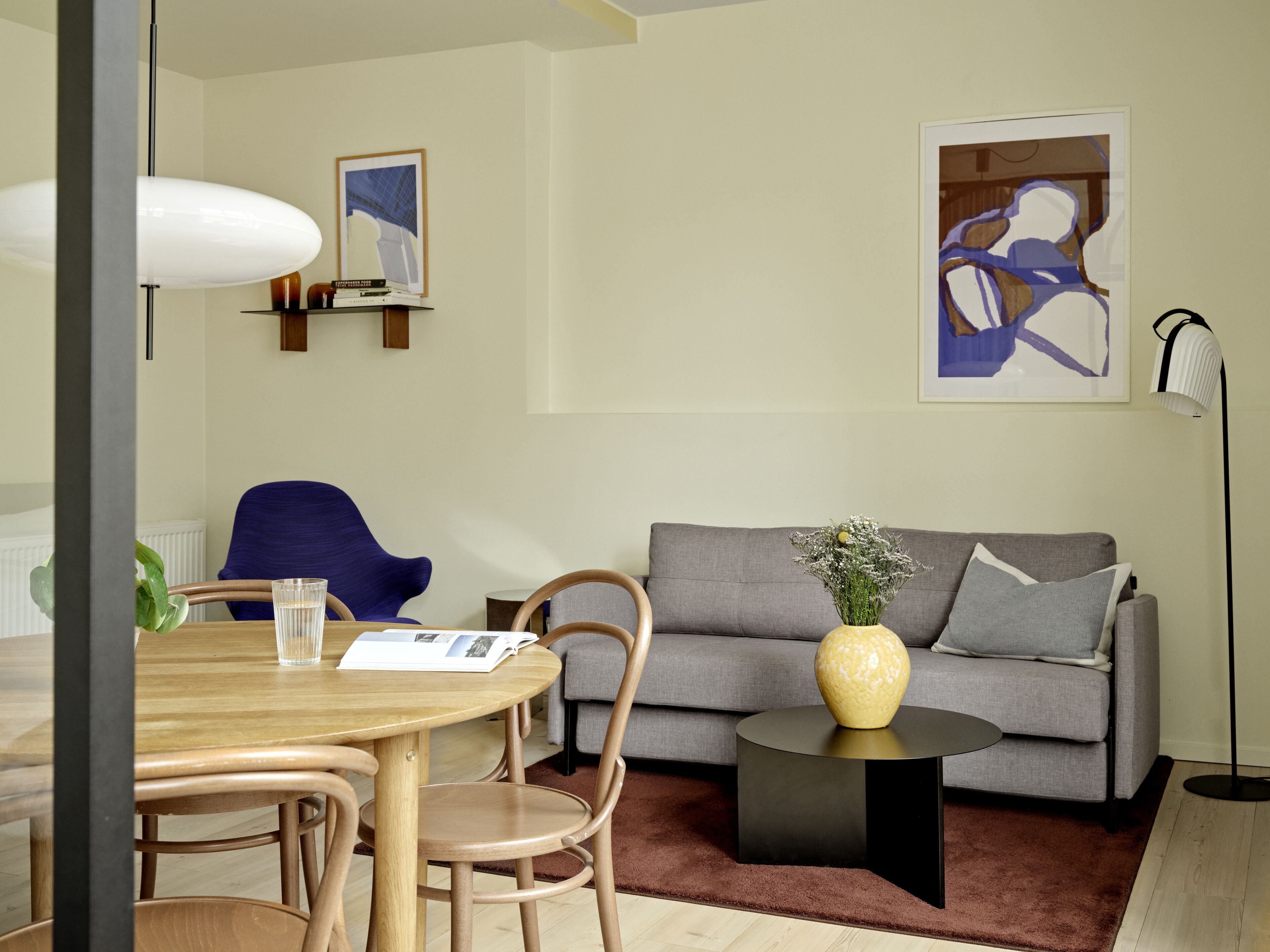 A modern living room with a grey sofa, a round wooden table, and a coffee table. There are two chairs, a floor lamp, and a framed picture on the wall.