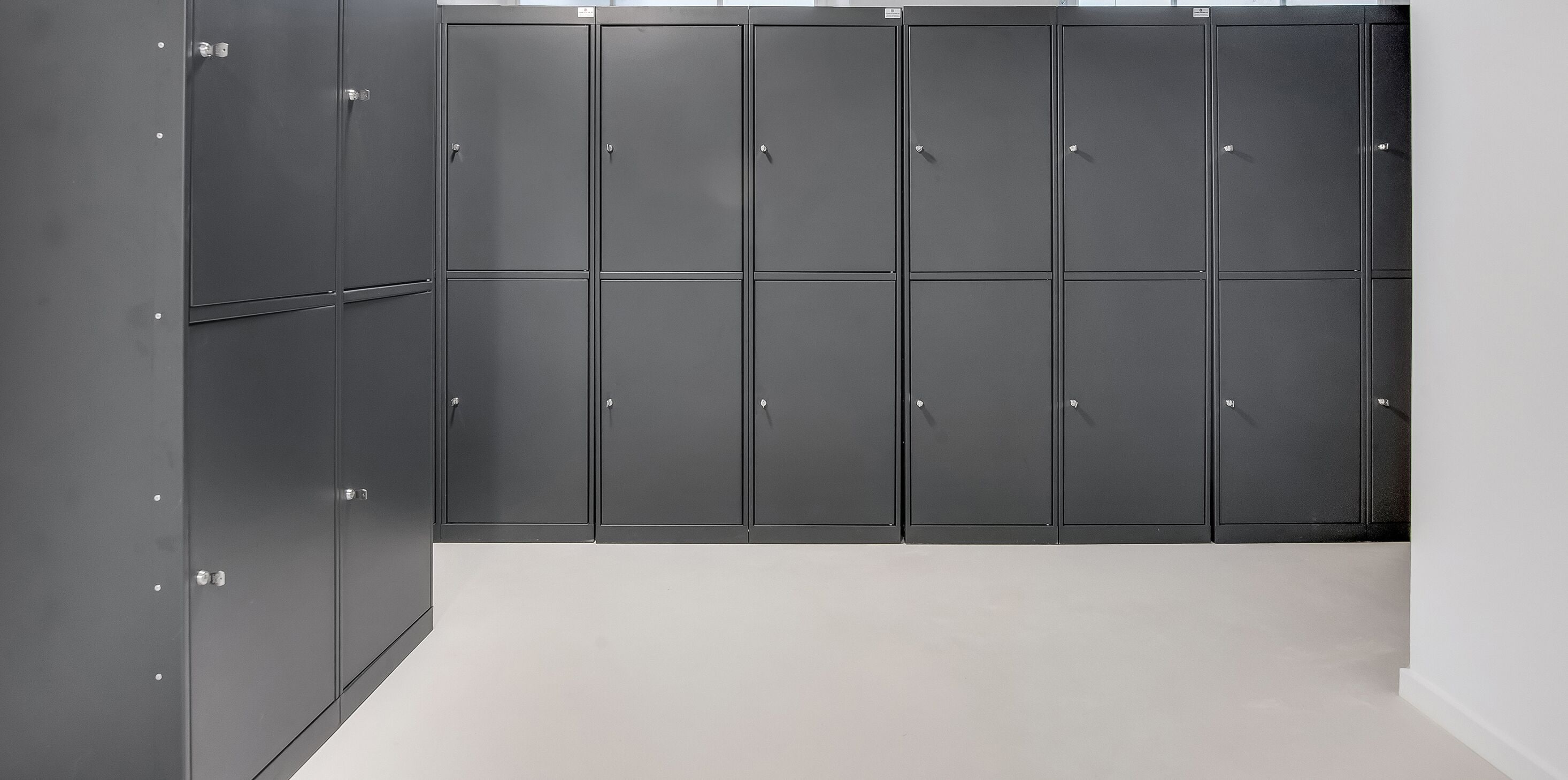 Une rangée de casiers gris fermés, dotés de poignées métalliques, est placée contre un mur uni dans une pièce bien éclairée.