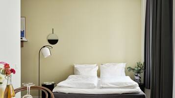 A hotel room with a bed, a bedside table with a lamp, a chair, and a table with a vase of flowers and a wine bottle.