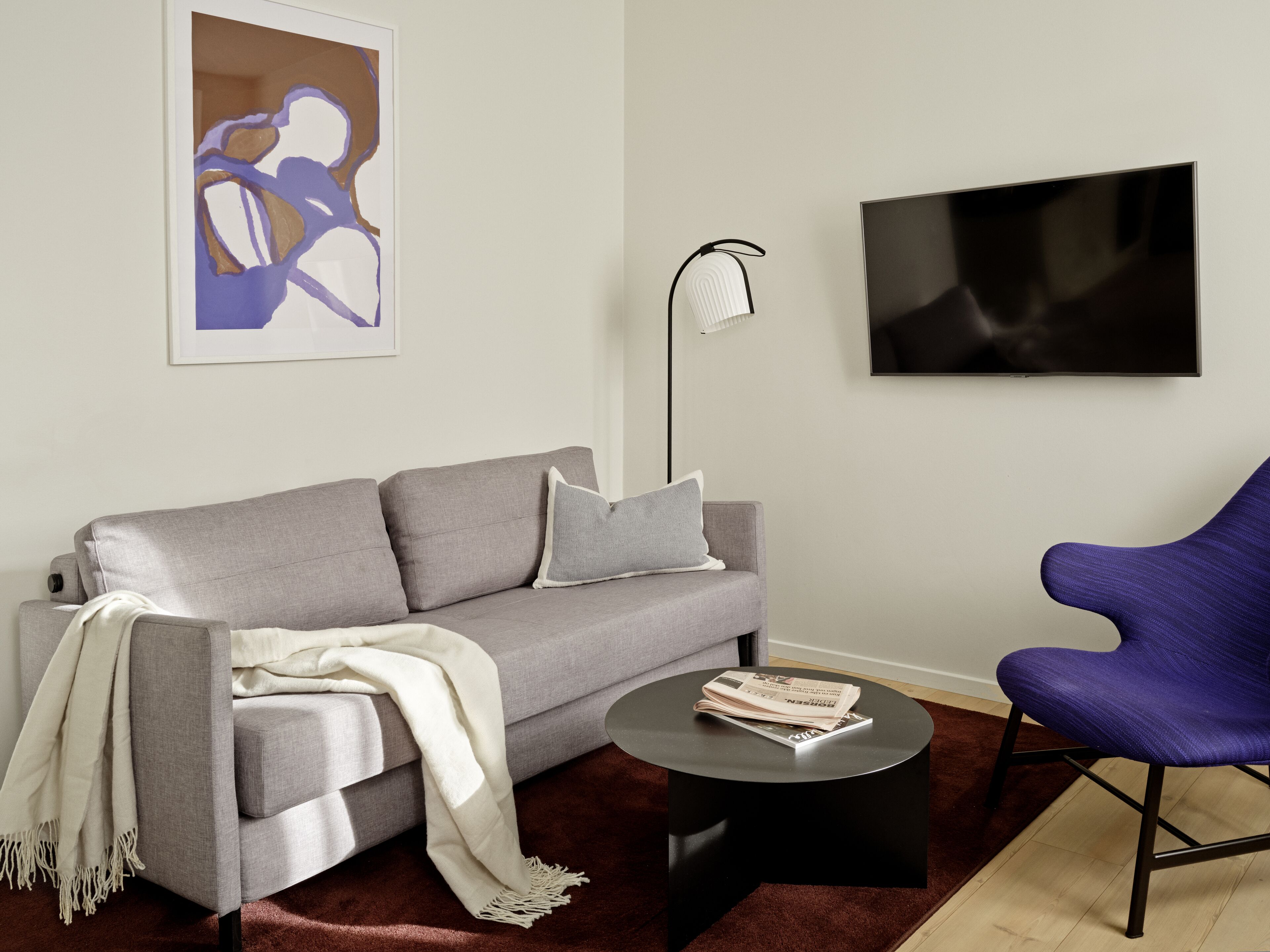 Un salon moderne avec un canapé gris, un plaid blanc, une table basse ronde noire, un fauteuil violet, un lampadaire et un tableau abstrait encadré.