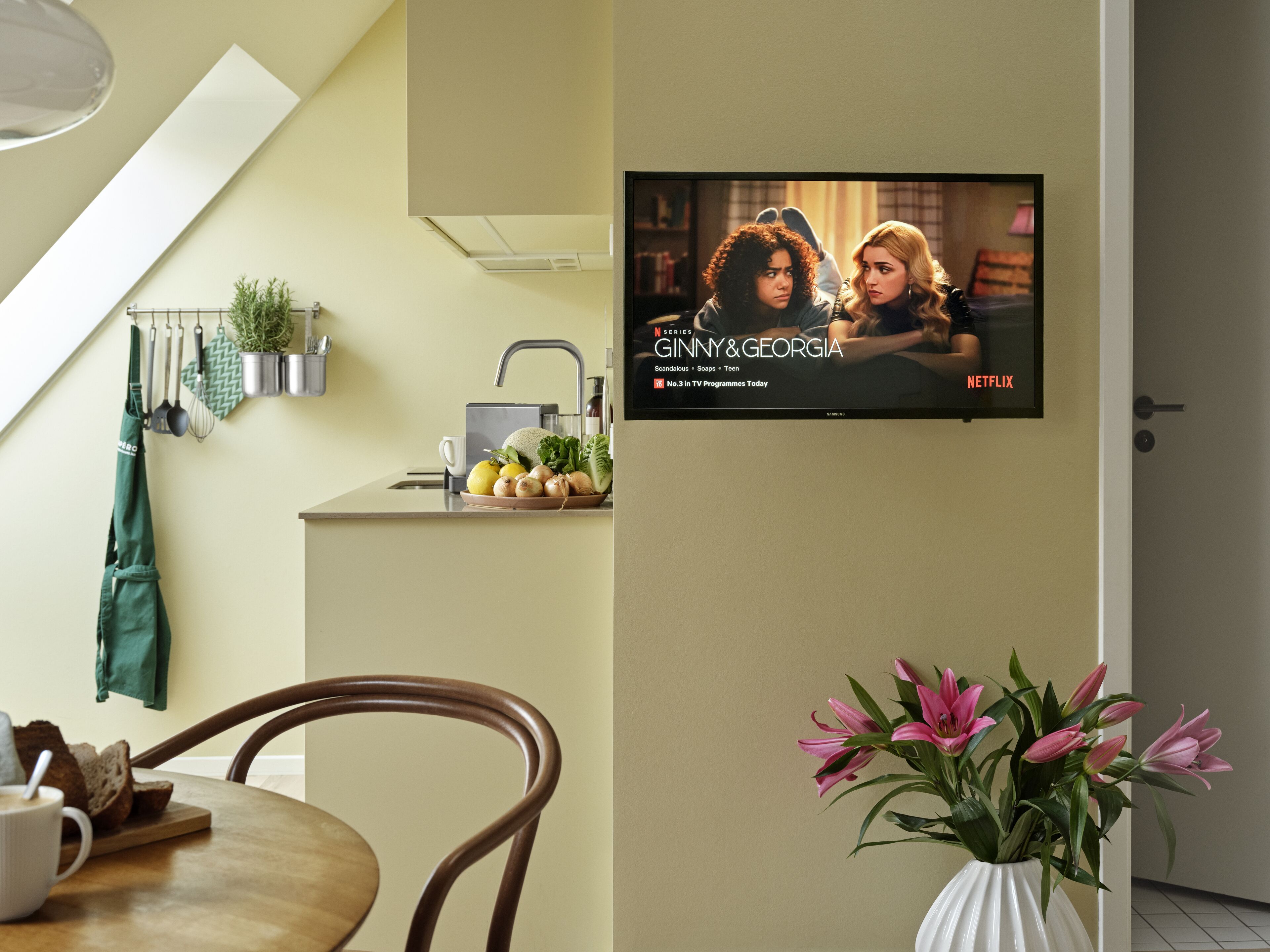 Une cuisine moderne avec un coin repas, un téléviseur diffusant la série « GINNY & GEORGIA » sur Netflix, et un vase rempli de fleurs roses.