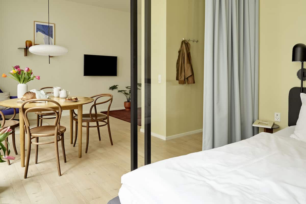 Une chambre d’hôtel moderne comprenant un lit, un coin repas avec une table et des chaises, un téléviseur fixé au mur et une plante en pot.