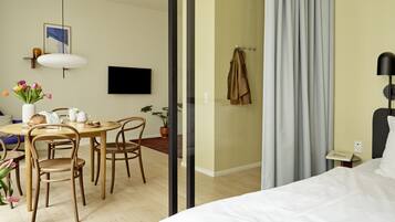 A modern hotel room with a bed, a dining area with a table and chairs, a TV mounted on the wall, and a potted plant.