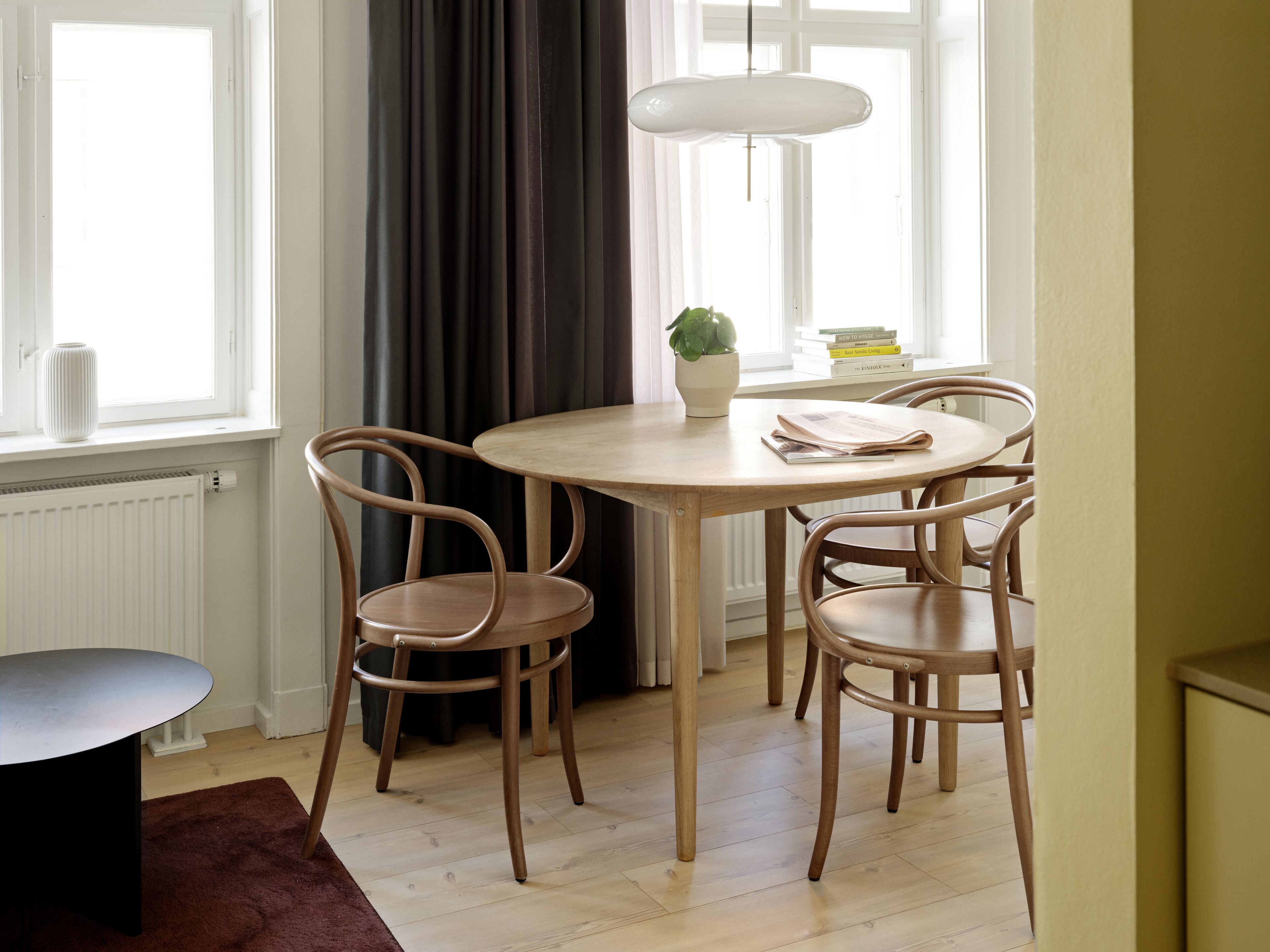 Un comedor con una mesa redonda de madera, cuatro sillas, una planta en maceta y una lámpara colgante.