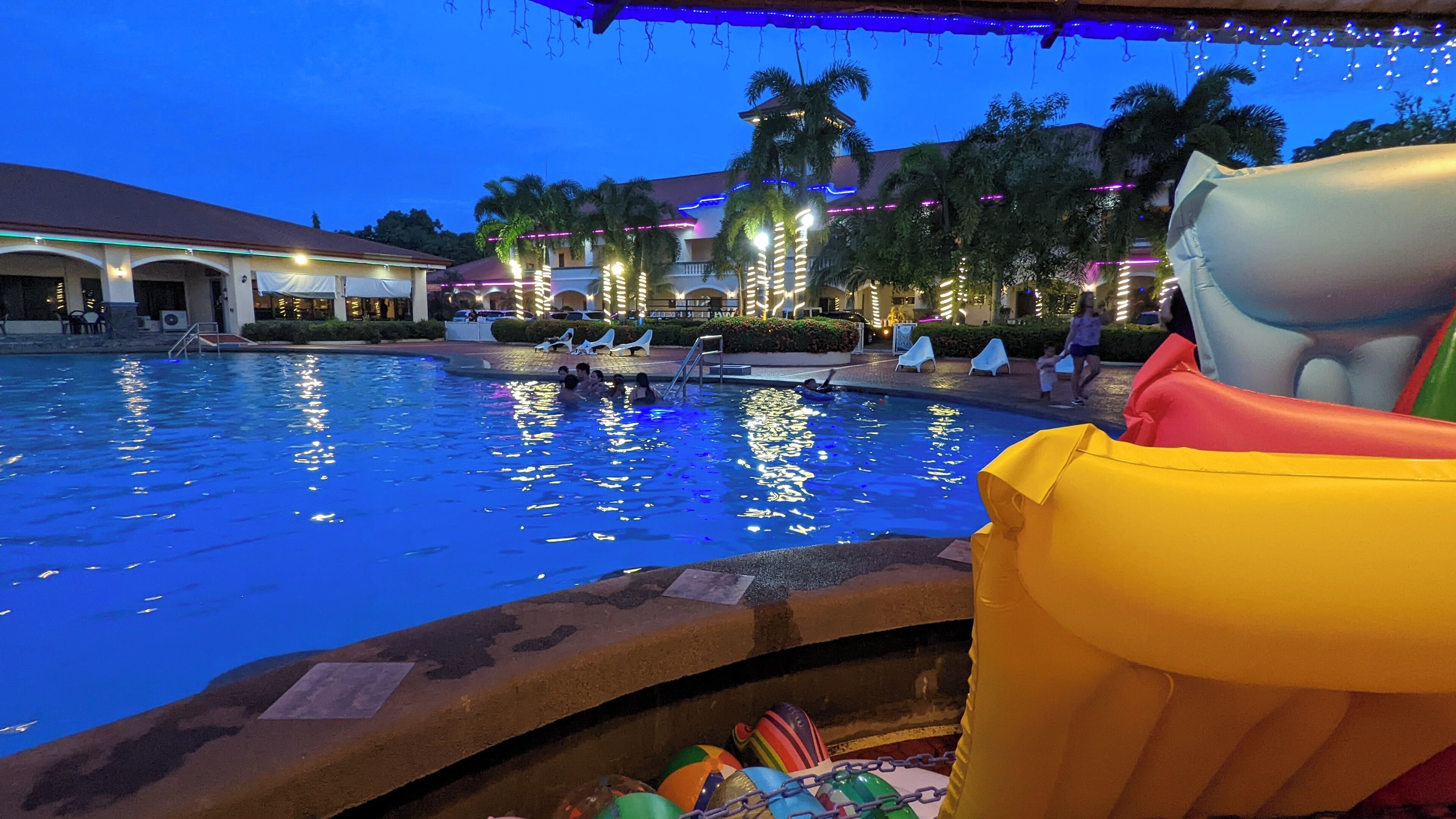 A swimming pool with people in the water, surrounded by palm trees and buildings with illuminated railings.