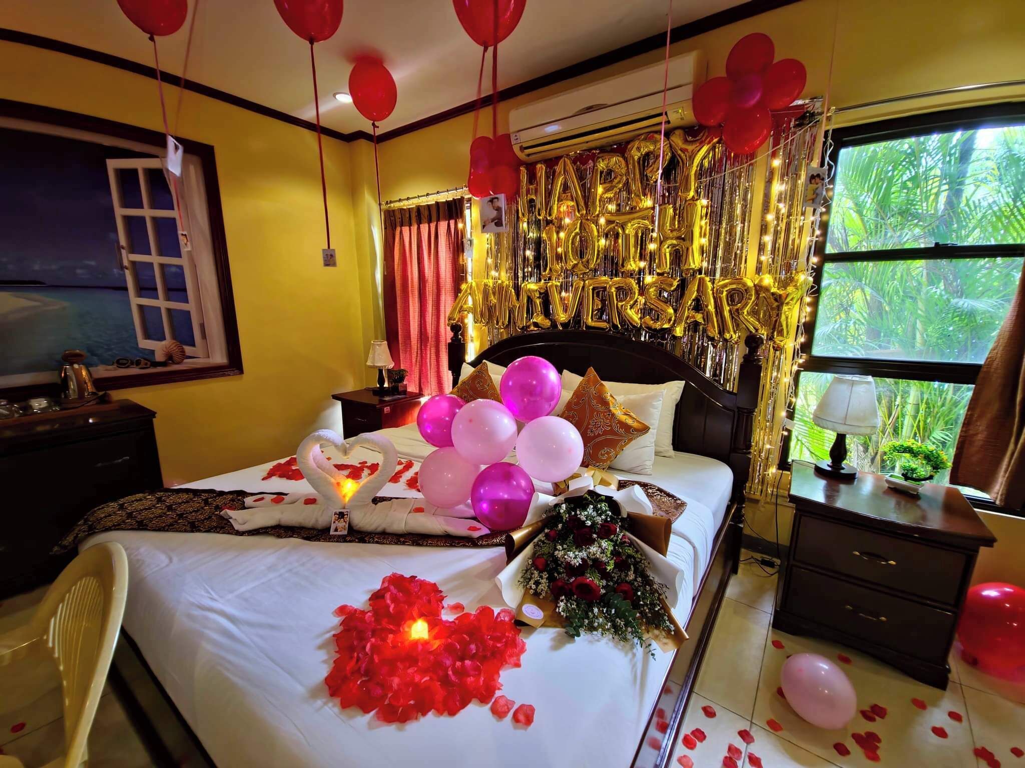 A hotel room with a bed adorned with balloons, a bouquet of flowers, and rose petals.
