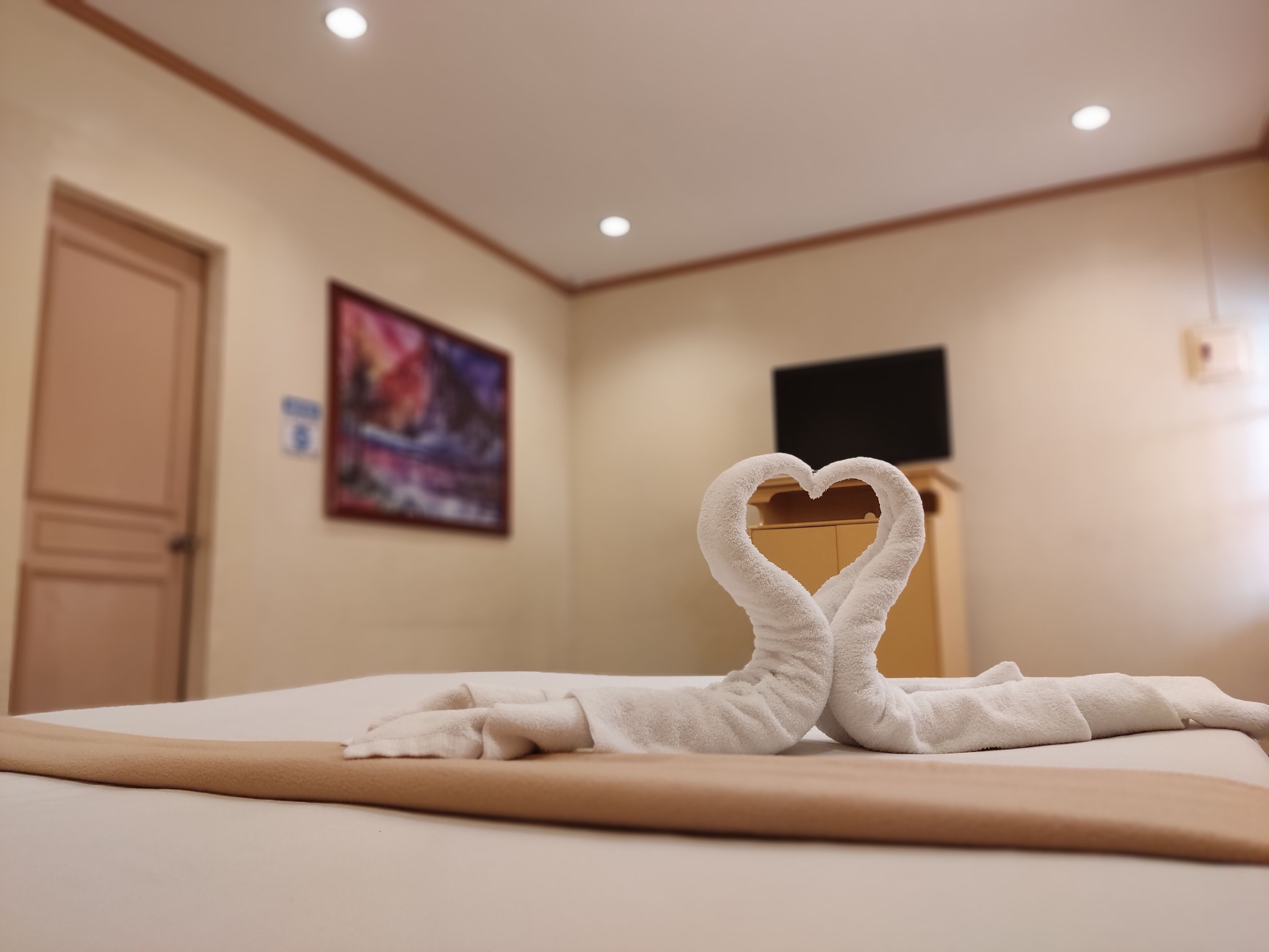 A hotel room with a bed, a heart-shaped towel arrangement, a painting on the wall, and a television on a stand.