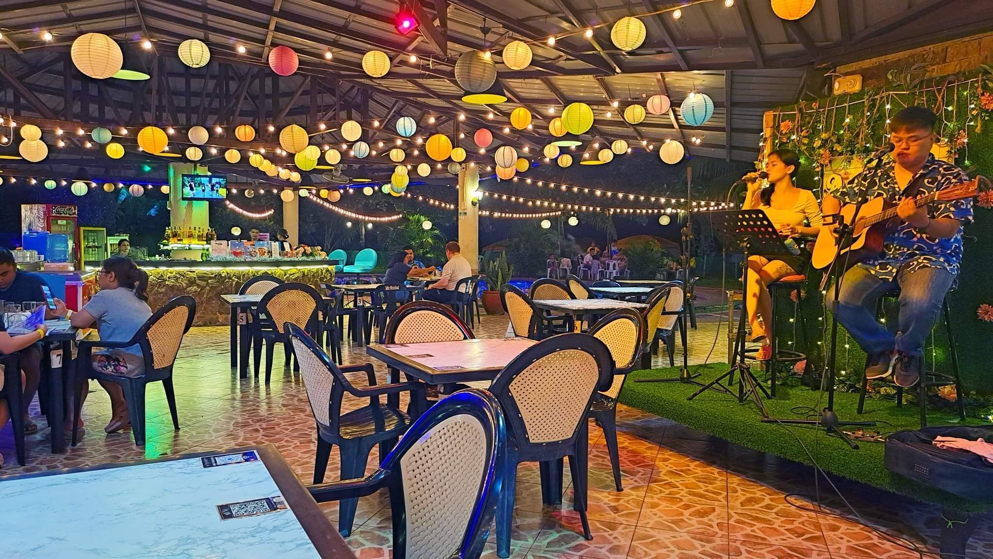 An outdoor dining area with tables and chairs, a stage with a performer, and string lights.