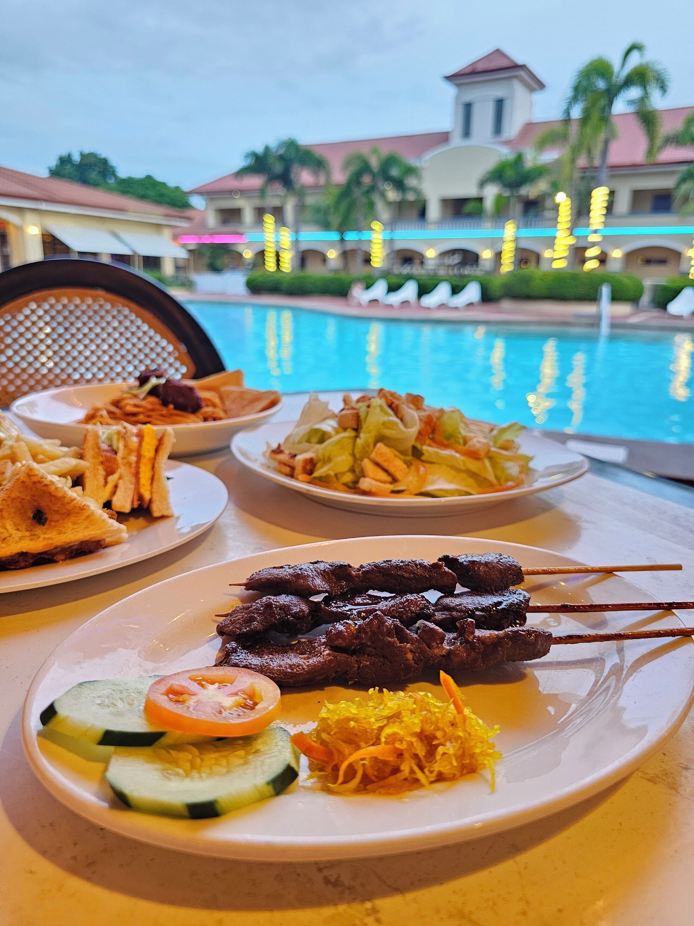 Una mesa de comedor con varios platos, incluyendo carne ensartada, papas fritas y un sándwich, con vista a una piscina y un edificio al fondo.